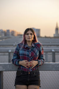 Portrait of young woman standing against railing during sunset