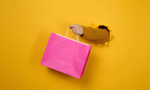 Female hand with a pink paper shopping bag sticking out of a torn hole in a yellow paper background. 