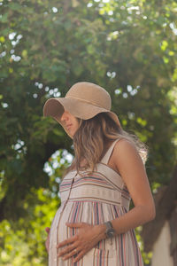 Low angle view of pregnant woman wearing hat while standing against trees