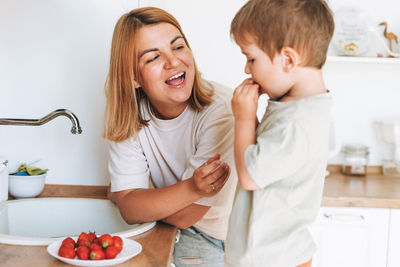 Young woman mother with her toddler boy son eat srawberries in kitchen at home