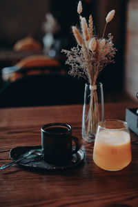 A cup of black coffee, orange juice and dried flowers in a glass vase standing on a wooden table 