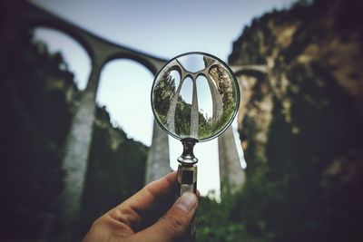 Cropped hand holding magnifying glass against viaduct bridge