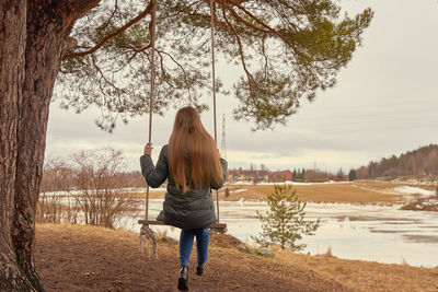 A young woman long blond hair on a wooden swings on a tree branch with icy river on the background..