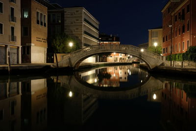 Illuminated bridge over river by buildings against sky at night