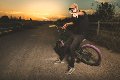 Portrait of man gesturing shaka sign while sitting on bicycle against sky during sunset