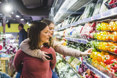 Cute daughter choosing bell peppers while being piggybacked by mother in store