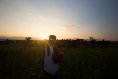Rear view of young woman holding flower while standing amidst plants on field against sky