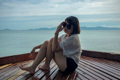 Young woman sitting on wooden chair by sea against sky