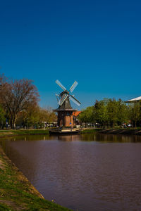 Traditional windmill by lake against clear blue sky