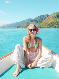 Portrait of woman wearing sunglasses while sitting in boat on sea against mountains and sky