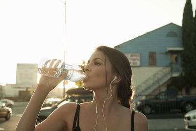 Young woman drinking water from bottle in city during sunset