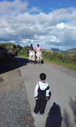 Rear view of boy looking at women riding horses on road