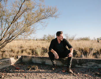 Full length of man sitting on retaining wall against bare tree and clear sky during sunny day