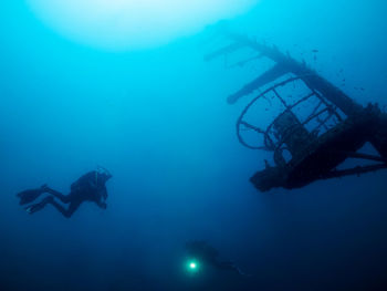 Professional divers with flashlight swimming near old sunken ship covered with moss on bottom of deep sea with clear water