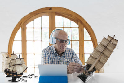 Senior man wearing headphones painting model ship on table with tablet