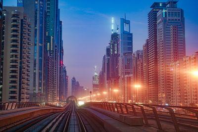 Illuminated railroad tracks amidst buildings in city against sky