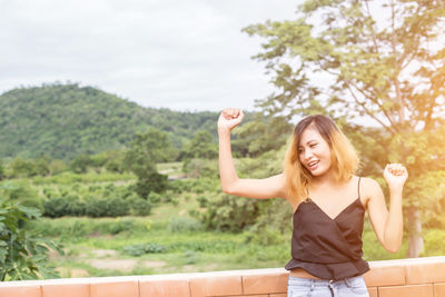 Happy young woman standing by plants against trees