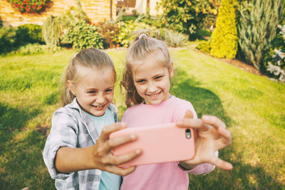 Two happy teenage girls laugh and take a selfie on a cell phone outdoors.