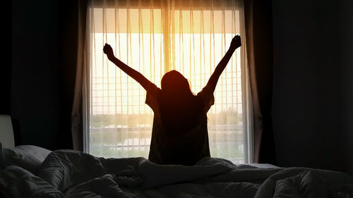 Rear view of silhouette woman stretching hands while sitting on bed at home