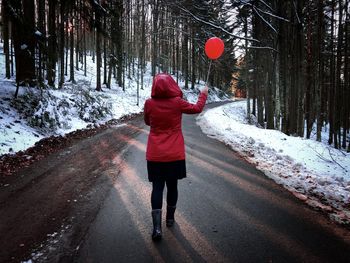 Rear view of woman with red jacket holding a red balloon and walking on road during winter