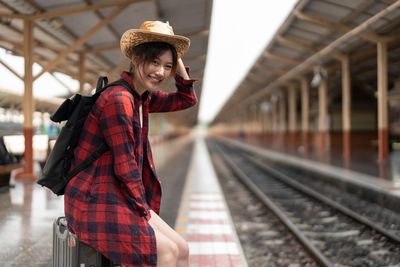 Happy young woman sitting on suitcase at railway station