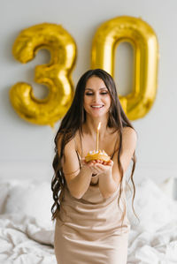 Happy woman blows out a candle on a birthday cake, sitting on a bed in a bedroom decorated 