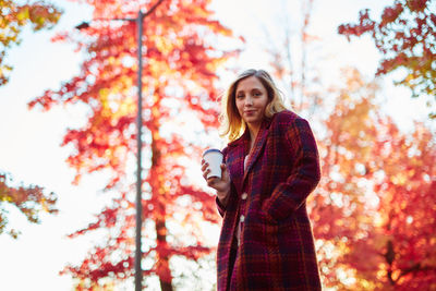 Young woman holding coffee while standing against autumn trees