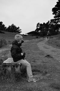 Side view of boy sitting on bench at grassy field against clear sky