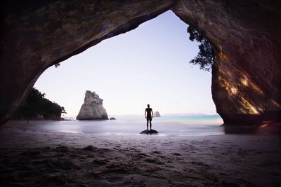 Man standing at beach seen through cave against sky