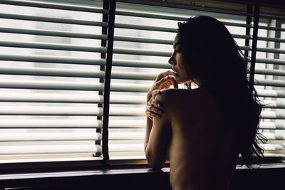 Shirtless woman looking through window at home