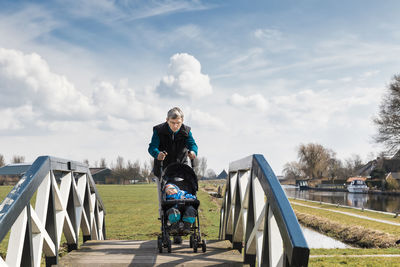 Man carrying granddaughter in baby stroller on footbridge at park against sky