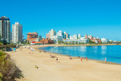 People on beach by buildings against clear blue sky