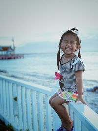 Smiling cute girl sitting on railing against sea at beach