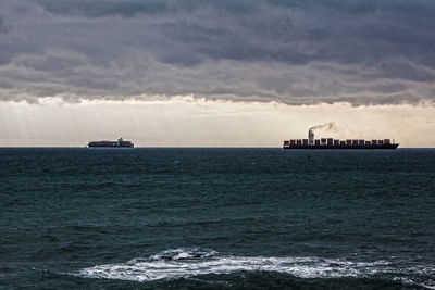 Distant view of container ships sailing on sea against cloudy sky