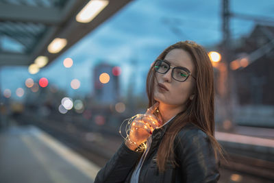 Portrait of young woman standing at railroad station platform during sunset