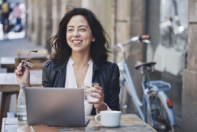 Cheerful woman looking away while sitting at sidewalk cafe with drinks and laptop