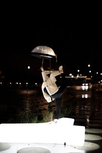 Woman holding umbrella standing on illuminated footpath at night