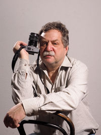 Portrait of mature man holding camera while sitting on chair against wall
