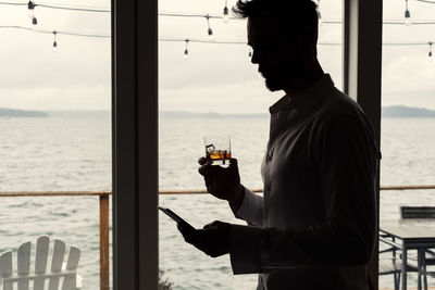 Man using mobile phone while drinking alcohol in glass by sea seen through glass window