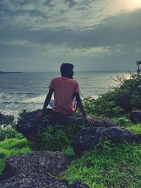 Rear view of woman sitting on rock by sea against sky