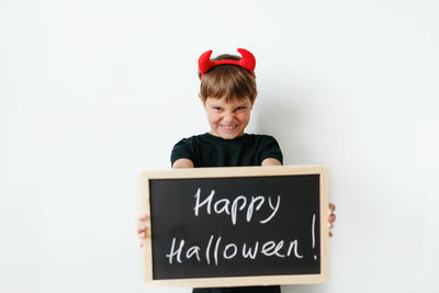 Little boy with red devil horns and black board with happy halloween text on white background