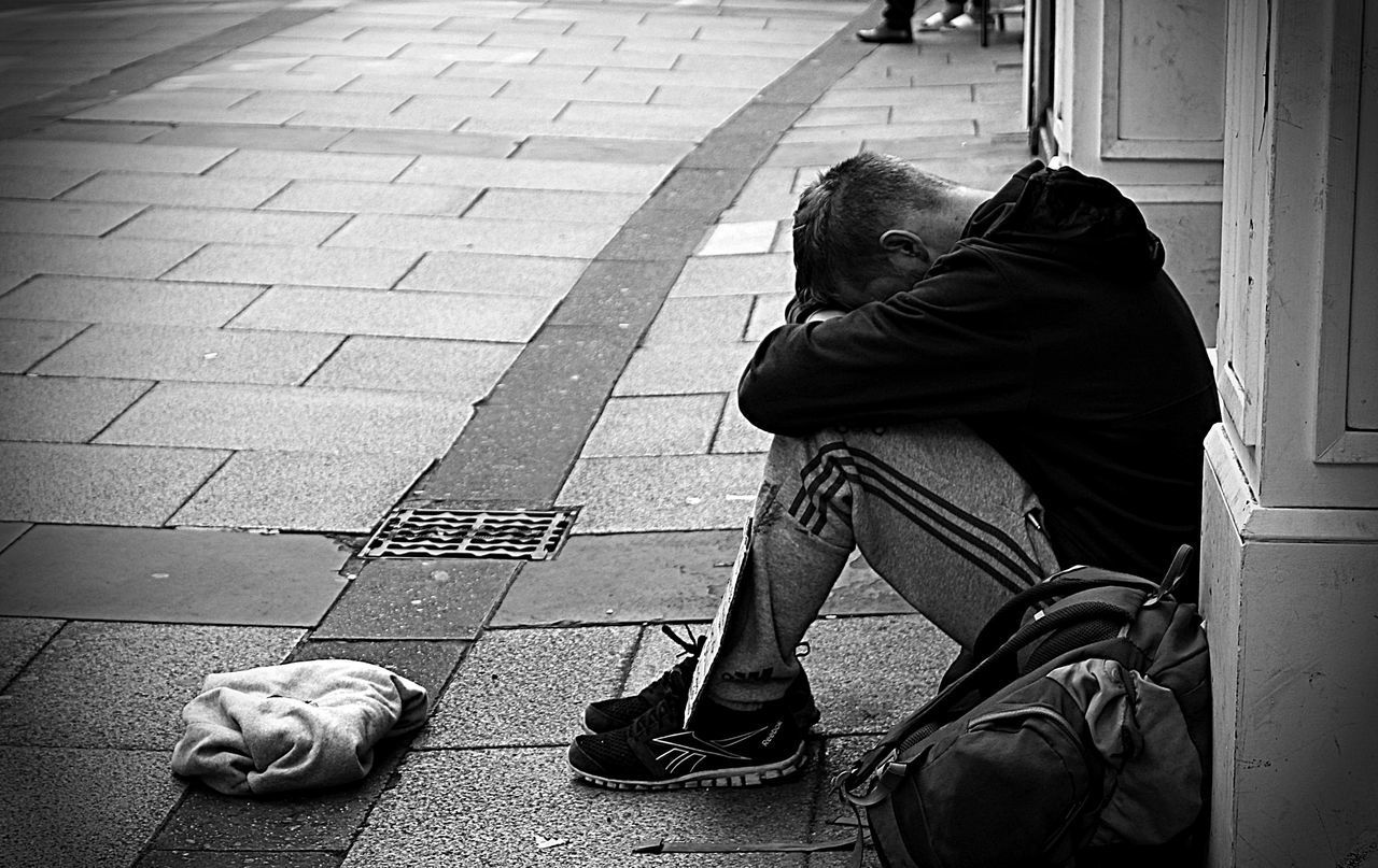 MAN SITTING ON FOOTPATH IN STREET