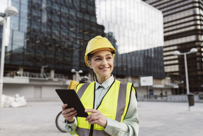 Happy engineer wearing hardhat standing with tablet pc outside building