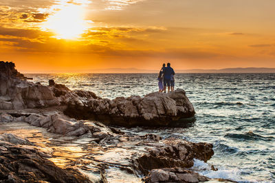 People on rock by sea against sky during sunset