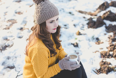 High angle view of young woman with coffee cup sitting on snow covered field during winter