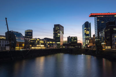 Illuminated buildings by river against sky at dusk