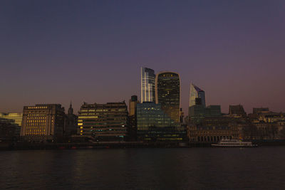 Modern buildings by river against sky during sunset