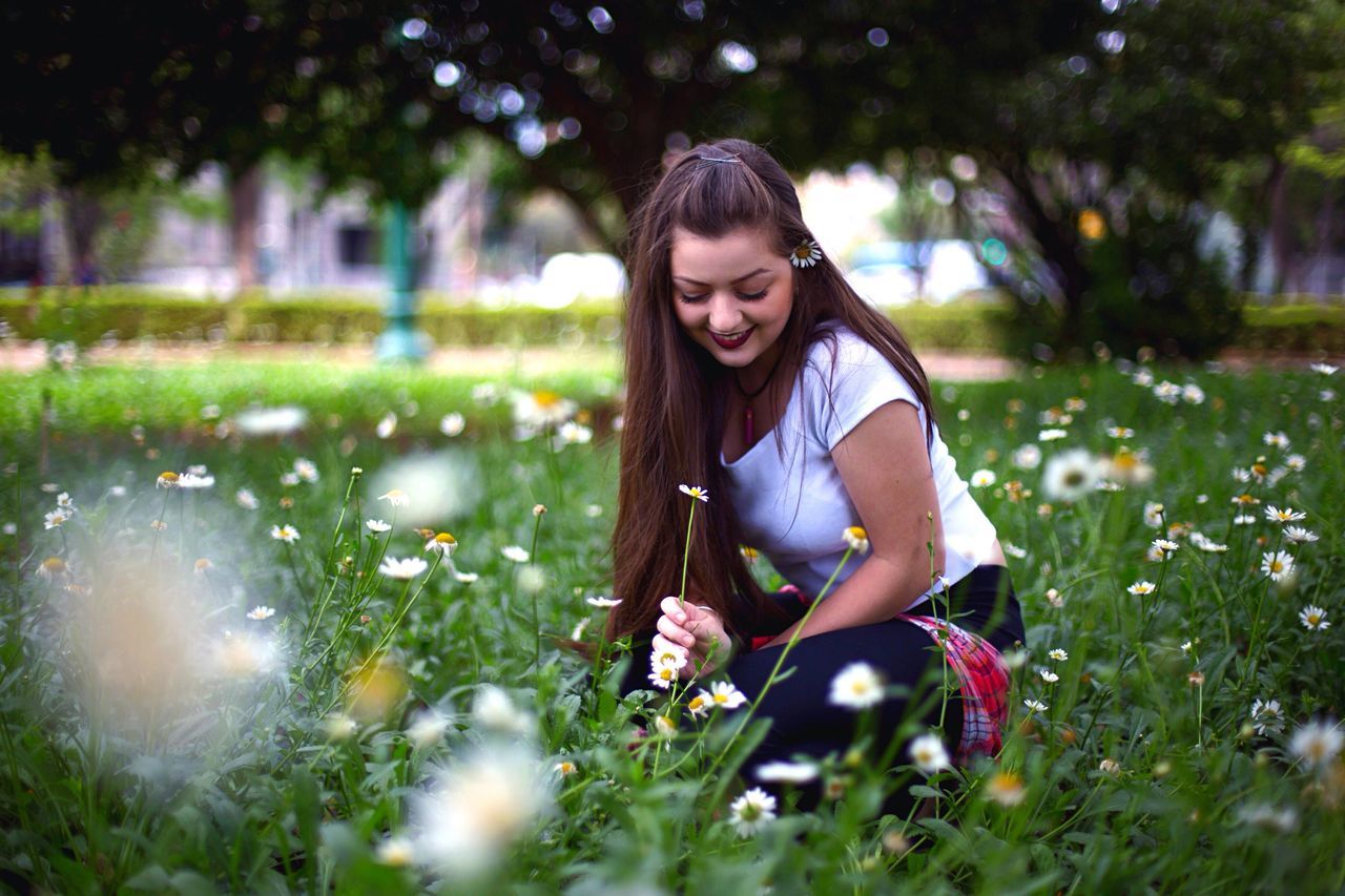 young adult, person, young women, tree, portrait, looking at camera, smiling, sitting, casual clothing, park - man made space, flower, park, selective focus, looking down, front view, day, confidence, nature, growth, beauty, focus on foreground, long hair, garden, outdoors