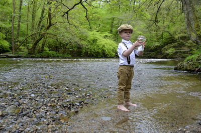 Full length of boy holding jar while standing by river in forest