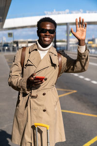 Black traveler man stands in airport terminal, holding mobile phone, calling taxi, raising hand